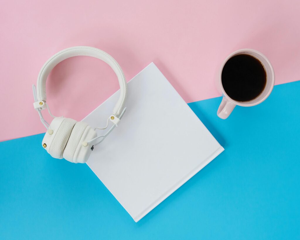 A minimalist flat lay featuring headphones, a coffee cup, and a blank notepad on a vibrant blue and pink background.